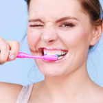Brunette woman winking while brushing her teeth with a pink toothbrush.