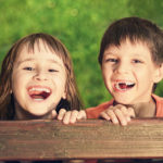 A young boy and girl smiling by a fence in the green grass.
