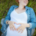 Happy pregnant woman in denim jacket lying on the green grass.