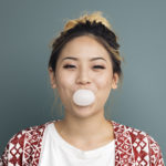 Asian woman with blonde hair and red jacket blowing a bubble of bubblegum